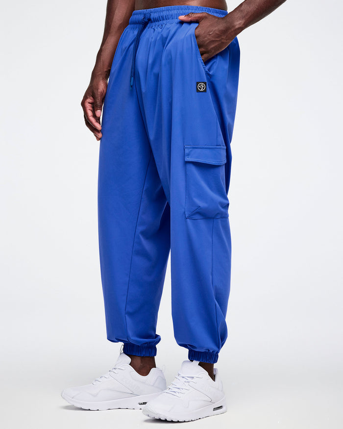 Male model wearing blue cargo pants with a brand logo just above his left pocket on a white background.