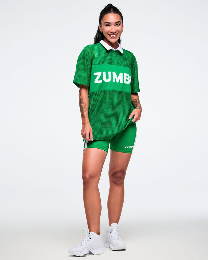 Full body view of a female model wearing a green mesh Zumba top with matching green biker shorts and white shoes.