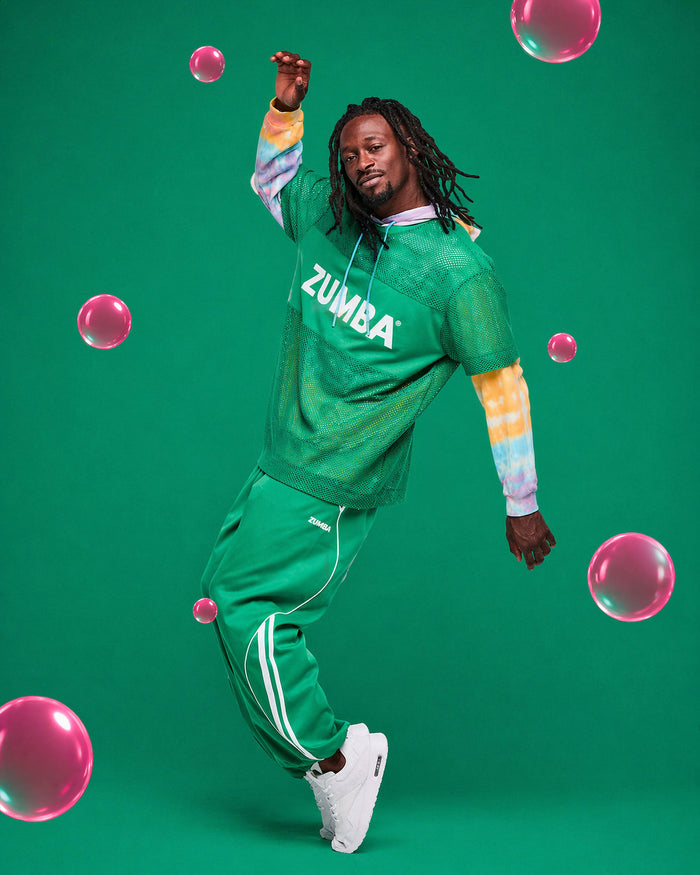 Man dancing in a green Zumba mesh top layered over a tie-dye hoodie, paired with green pants and white shoes, set against a green background with pink bubbles.