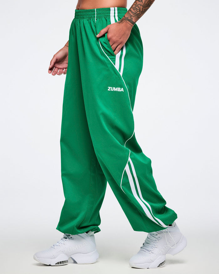 Female model with hands in pockets of the Zumba curvy track pants in green with white stripes on a white background.