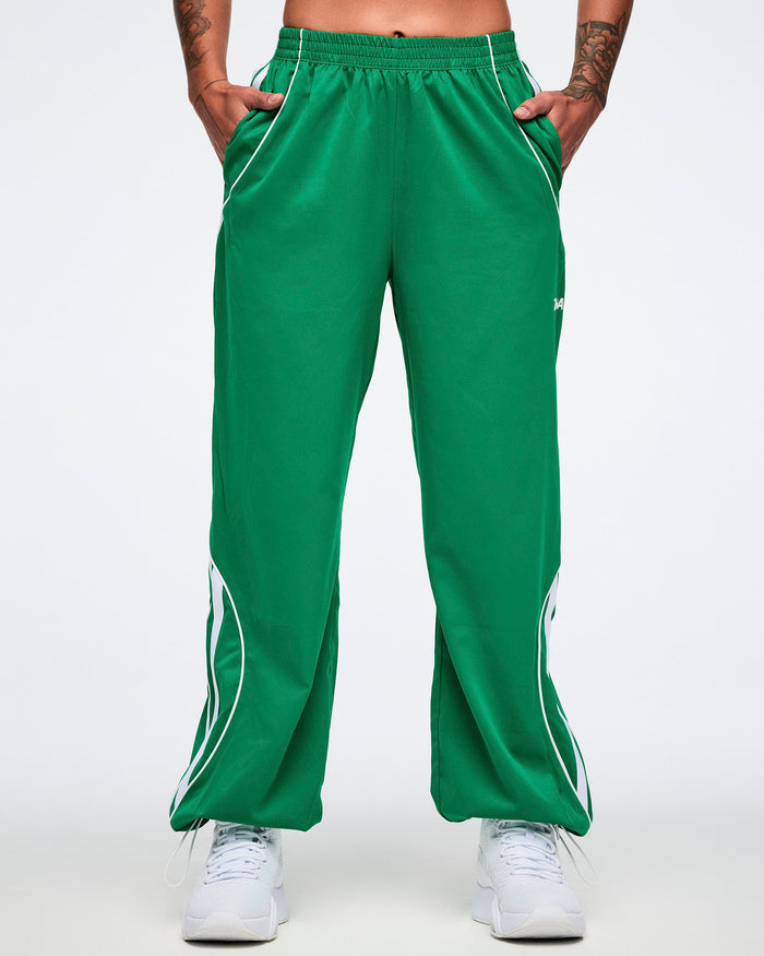 Female model with hands in her pockets of the green Zumba curvy track pants in with white stripes on a white background.