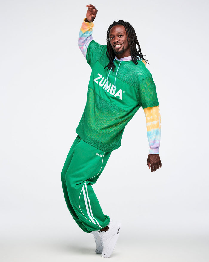Male model on  his toes dancing while wearing a green Zumba mesh top, a tie dye hoodie underneath and green curvy track pants.