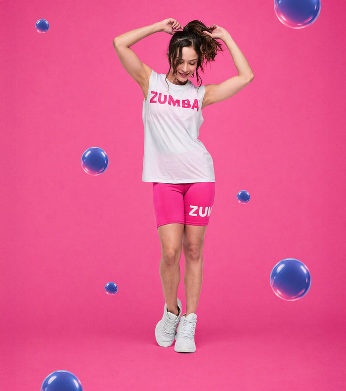 Female model dancing in a mesh crew neck muscle tank featuring pink "Zumba" lettering, paired with pink shorts and white shoes, posed against a pink backdrop with blue bubbles.