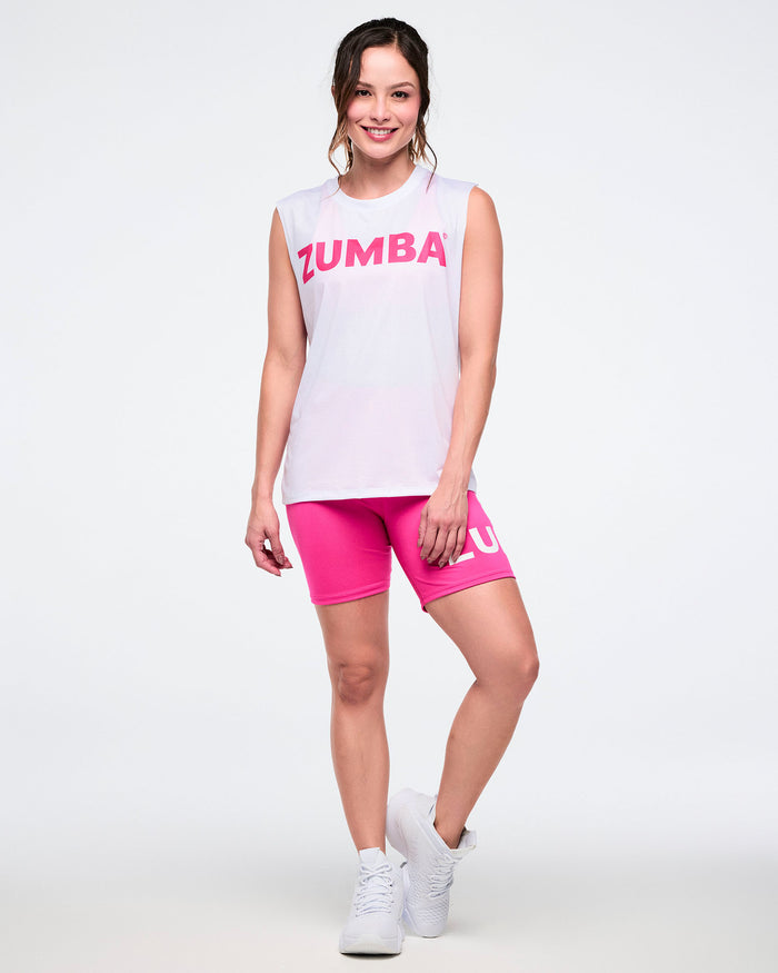 Female model wearing a mesh crew neck muscle tank featuring pink "Zumba" lettering, paired with pink shorts and white shoes against a white background.