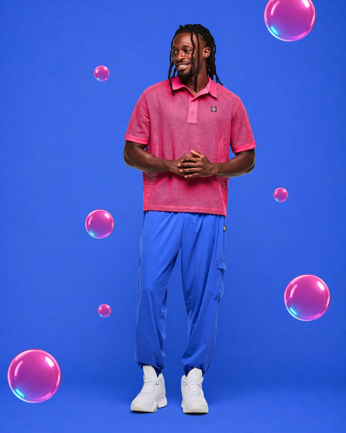 Man wearing a pink mesh polo shirt and blue pants standing against a blue background with pink bubbles.