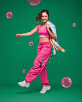 Female model in pink Zumba racerback and matching cargo pants with a tie-dye hoodie in her hand dancing on a green background with pink bubbles.