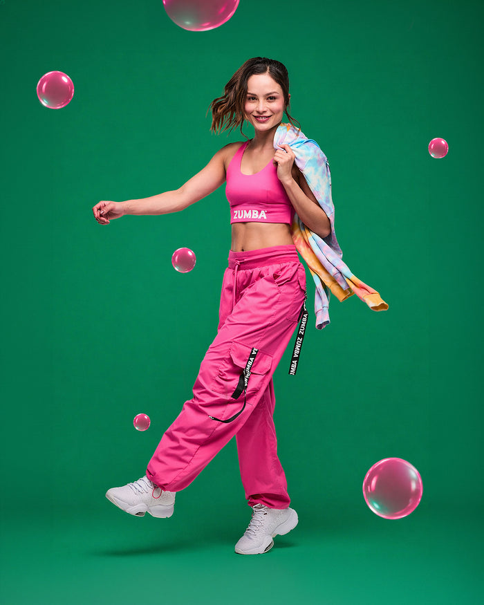 Female model in pink Zumba racerback and matching cargo pants with a tie-dye hoodie in her hand dancing on a green background with pink bubbles.