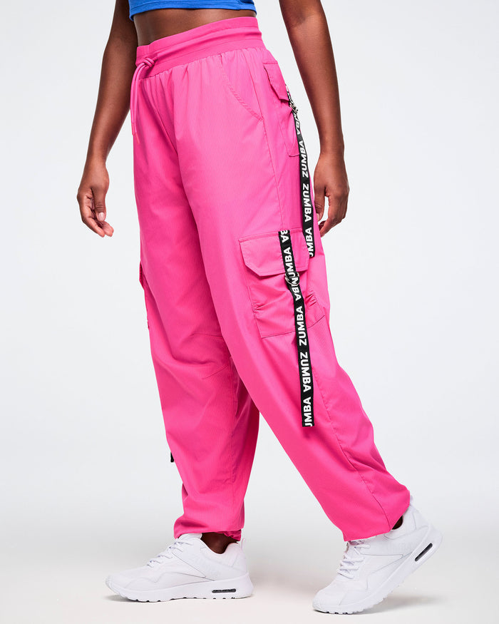 Female model wearing bright pink cargo pants accented with black pocket ribbons featuring white Zumba lettering, styled with white shoes.