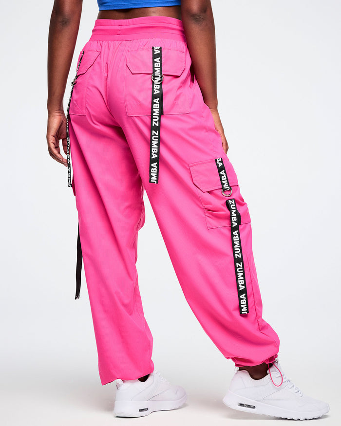 Back view of a female model wearing  bright pink cargo pants accented with black pocket ribbons featuring white Zumba lettering, styled with white shoes.
