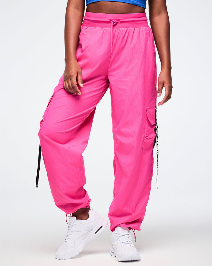 Female model wearing pink cargo pants accented with black pocket ribbons featuring white Zumba lettering, styled with white shoes against a white background.