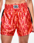 Female model wearing orange boxing shorts with an allover red “ZUMBA” monogram, a central ZW logo on the elastic waistband, and black side stripes.