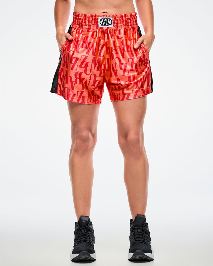 Front view of a female model with both hands in her pockets, wearing orange boxing shorts with an allover red “ZUMBA” monogram, paired with black-and-white shoes.