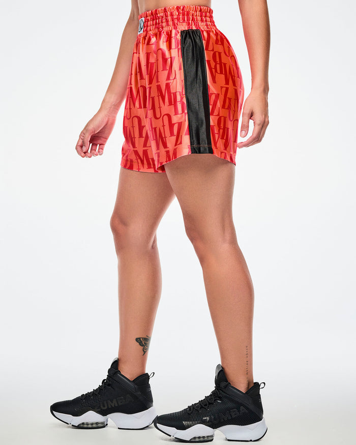 Side view of a female model wearing orange boxing shorts with an allover red “ZUMBA” monogram, paired with black-and-white shoes.