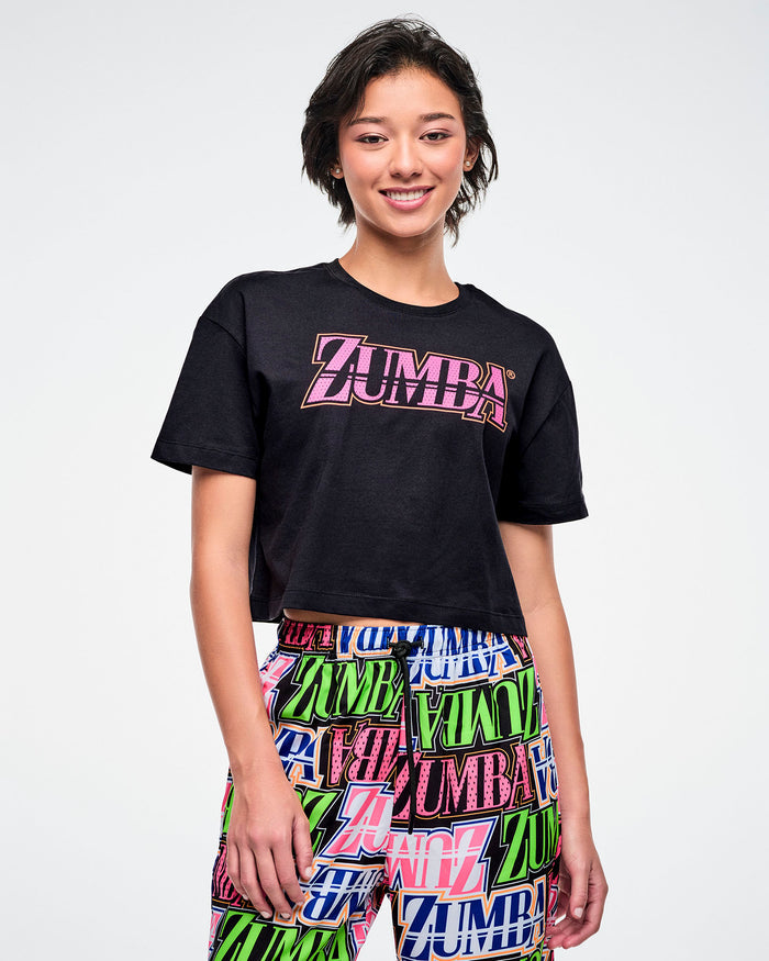 Female model in a black Zumba T-shirt with pink “ZUMBA” across the chest, paired with multicolored patterned Zumba pants.