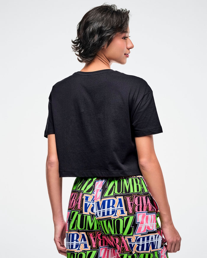 Back view of a female model in a black Zumba T-shirt, paired with multicolored patterned Zumba pants.