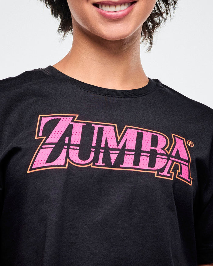 Close-up view of the female model in a black Zumba T-shirt with pink “ZUMBA” across the chest in a mesh-like pattern.