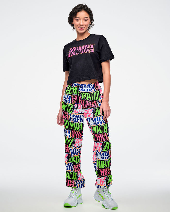 Female model in a black Zumba T-shirt with pink “ZUMBA” across the chest, paired with multicolored patterned Zumba pants and green and white shoes.