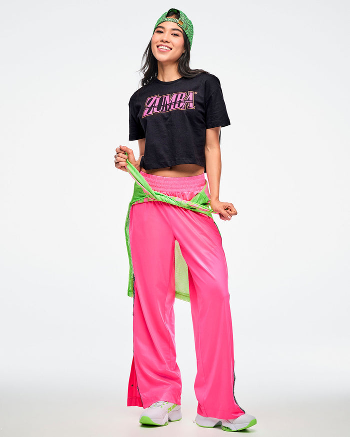Female model wearing a backward green Zumba hat, a black Zumba T-shirt with pink “ZUMBA” across the chest, pink Zumba pants, and green-and-white shoes, with a green shirt tied around her waist.