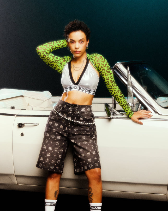 Female model posing in a silver V-neck bra with black trim, a green leopard-print long-sleeve top covering her arms, and denim jorts, leaning against a white vintage convertible.