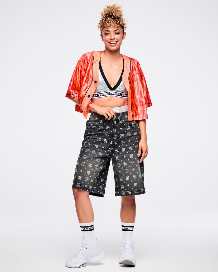 Female model wearing an open red button-up top over a silver bra, pulling up black denim jorts with one hand that features ZW, “01,” stars, dots, and Zumbito motifs, paired with black-and-white socks and shoes.