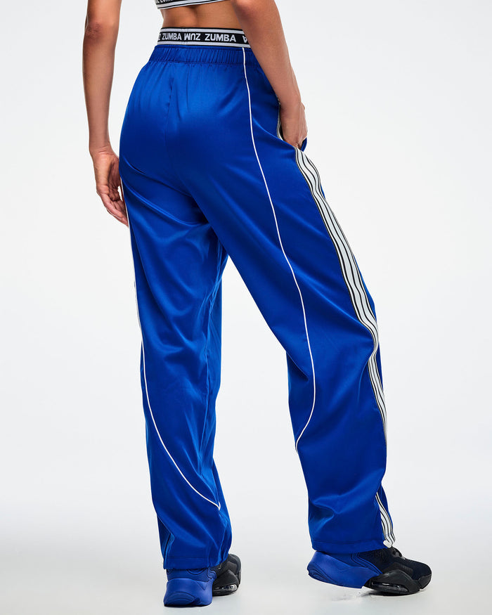 Back view of a female model with hand in her pocket, wearing blue woven pants with white stripes paired with blue and black shoes.