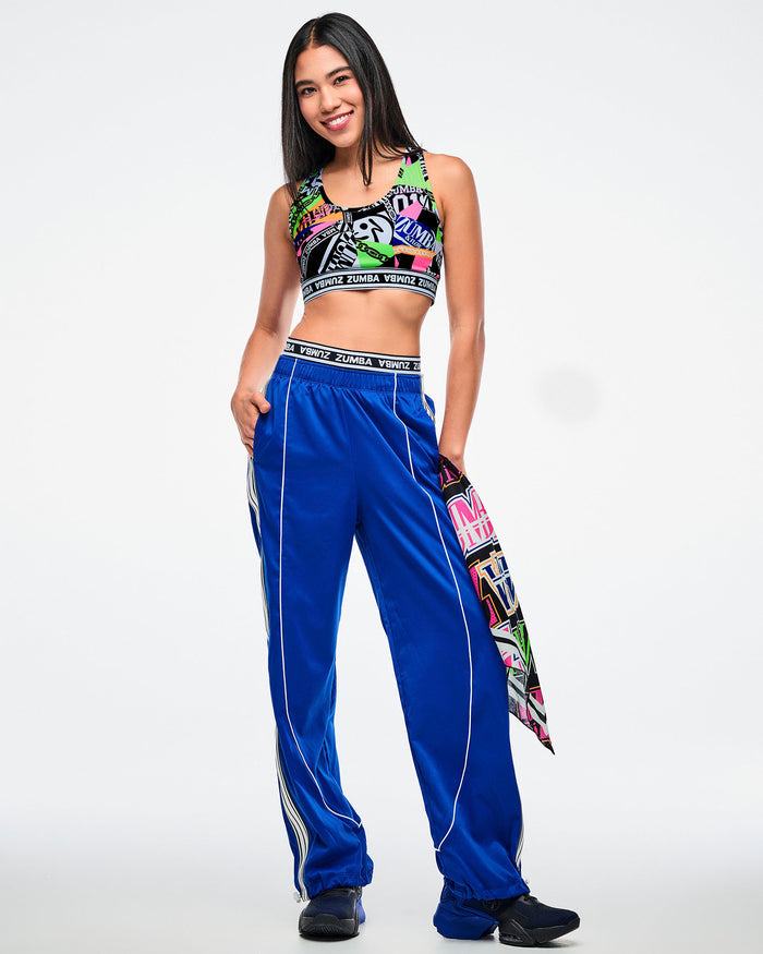 Female model in a colorful sports bra and blue woven pants with white side stripes, with a multicolor bandana hanging from her pocket and paired with blue and black shoes.