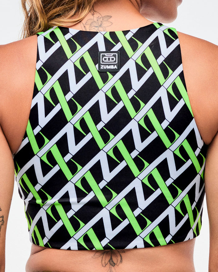 Back view of a female model wearing black based crop top with an allover white and green “Z” crisscross pattern with a black and white Zumba patch on the top.