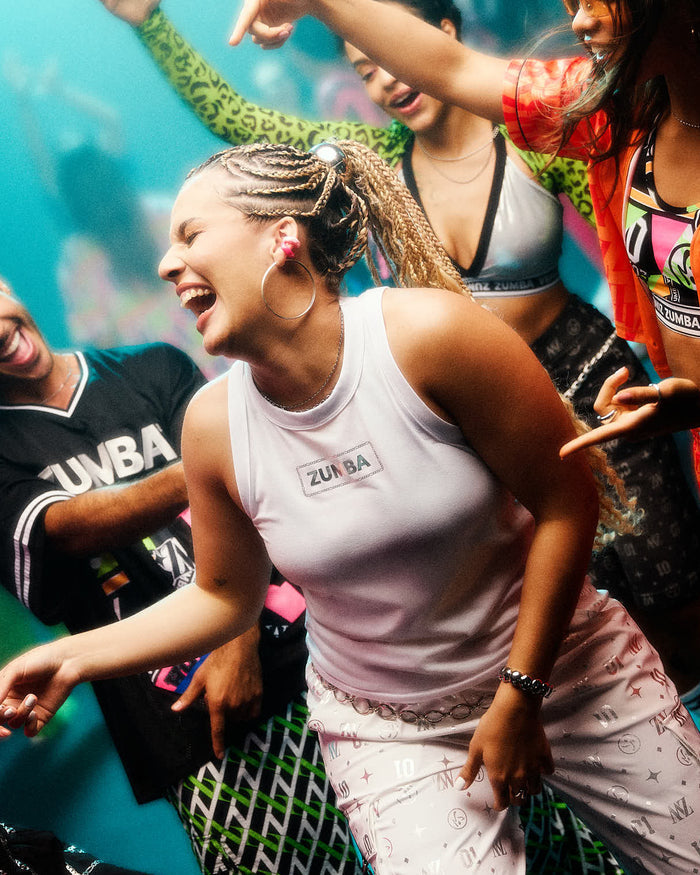 Female model dancing energetically in a white ribbed tank top with a silver 'Zumba' logo on the chest and matching patterned pants, surrounded by other people at a party.