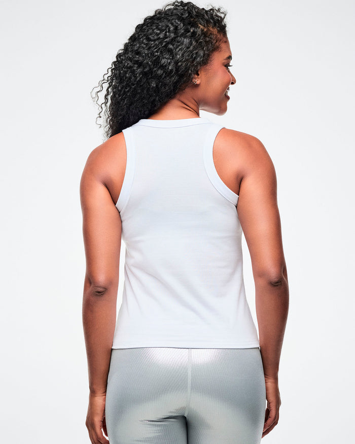 Back view of a female model wearing a white ribbed tank top with a silver 'Zumba' logo on the chest, paired with silver biker shorts on a white background.