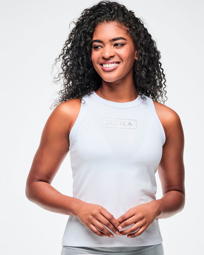 Close view of a female model wearing a white ribbed tank top with a silver 'Zumba' logo on the chest, paired with silver biker shorts.