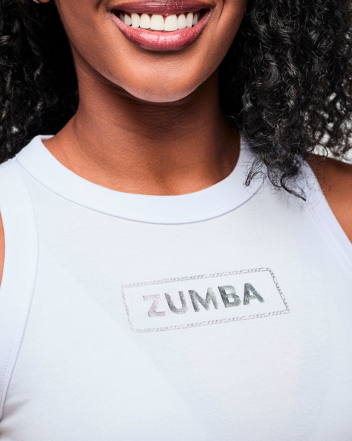Close-up detailed view of a female model wearing a white ribbed tank top with a detailed view of the silver 'Zumba' logo on the chest.