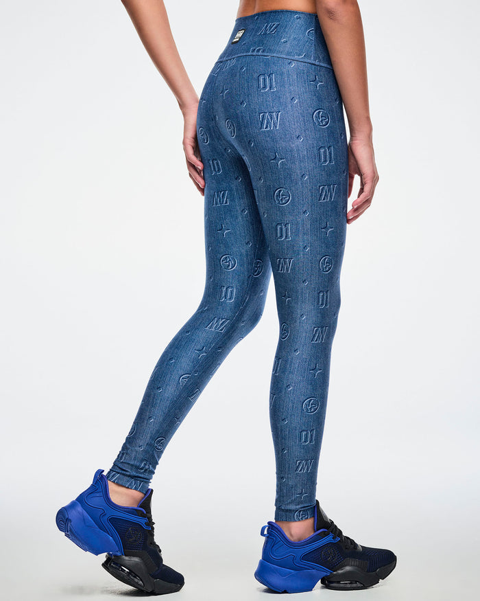 Back view of a female model wearing denim-print leggings with “ZW, 01, dots, Zumbito” and a rear patch, paired with blue-black shoes.