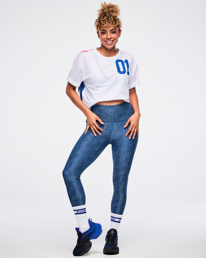 Female model wears a full Zumba outfit: a white mesh crop top, blue denim-patterned leggings, white-blue socks, and blue-black shoes against a white background.