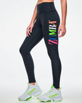 Female model wearing black Zumba leggings with colorful “ZUMBA” text down the left leg and paired with green-and-white shoes.