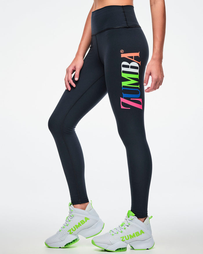 Female model wearing black Zumba leggings with colorful “ZUMBA” text down the left leg and paired with green-and-white shoes.