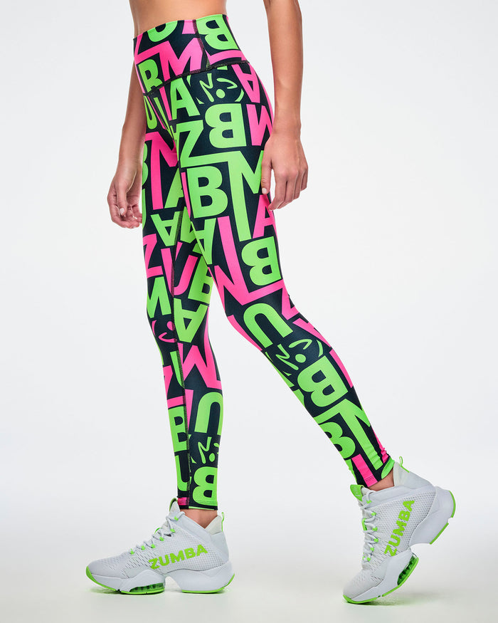 Female model wearing leggings with a black base and green and pink “ZUMBA” patterned lettering paired with green and white shoes.