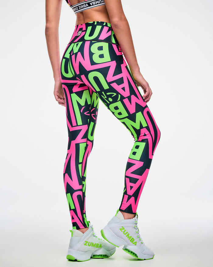 Back view of a female model wearing leggings with a black base and green and pink “ZUMBA” patterned lettering on a white background paired with green and white shoes.