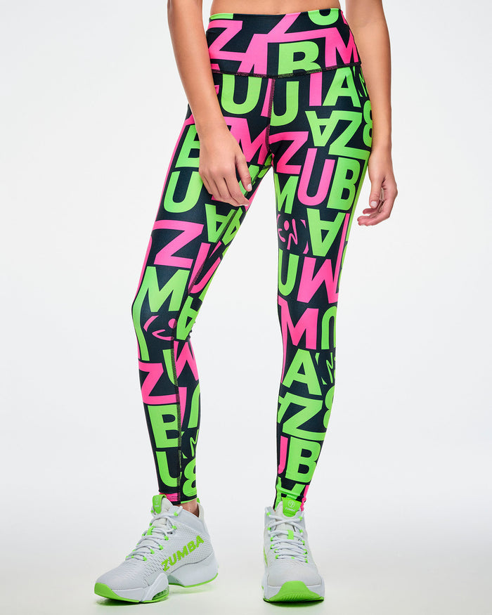 Front view of a female model wearing leggings with a black base and green and pink “ZUMBA” patterned lettering on a white background paired with green and white shoes.