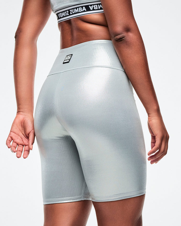 Back view of silver ribbed biker shorts with a black‑and‑white Zumba patch centered on the rear waistband.