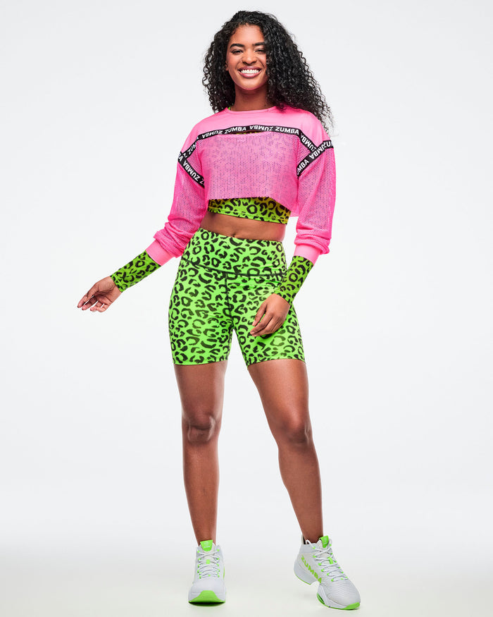Female model in a pink long‑sleeve cropped top layered over a green leopard‑print crop top, matched with green biker shorts and white‑and‑green sneakers.