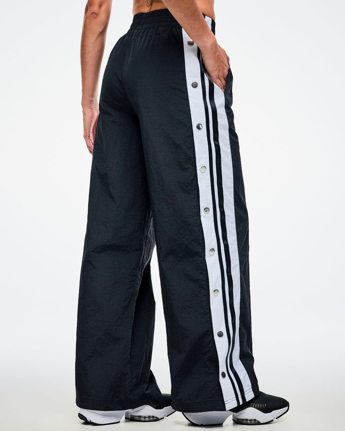 Back view of a female model wearing black pants with white stripes on the side with side snaps and paired with black and white shoes.