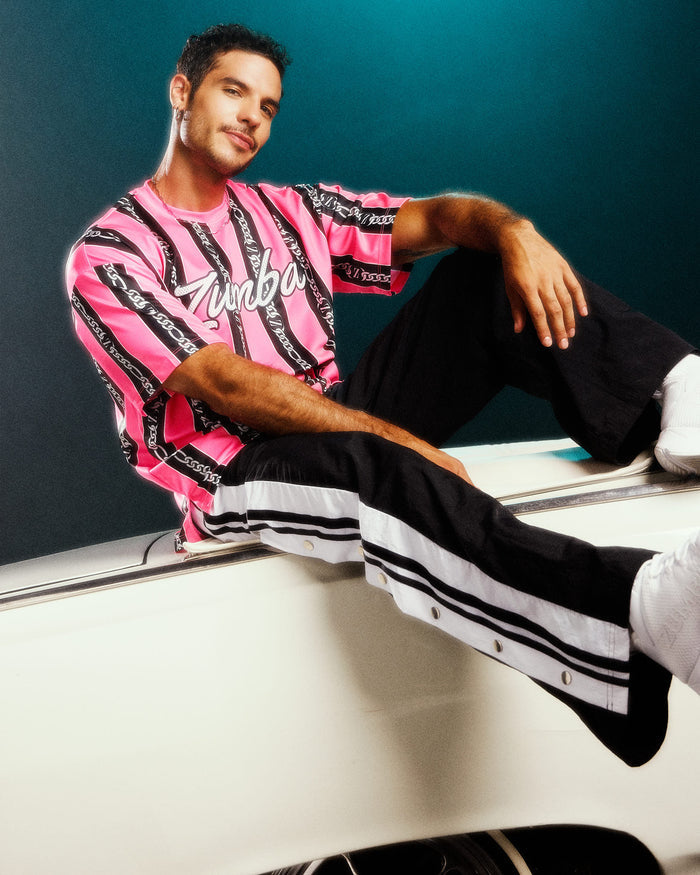 Male model leaning on a white car and wearing a pink and black striped top with 'Zumba' text paired with black pants with white stripes and white shoes.