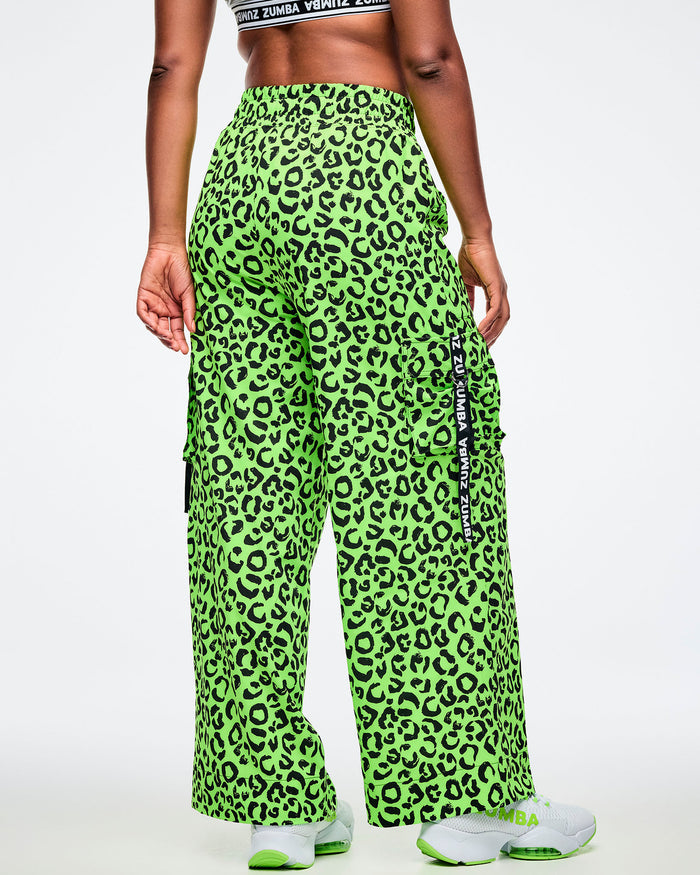 Back view of a female model wearing a high-waisted neon green cargo pants with black leopard print, elastic waistband, and side cargo pocket featuring a vertical black tape that reads “Zumba.” Styled with white athletic sneakers accented in matching neon green all on a white background.