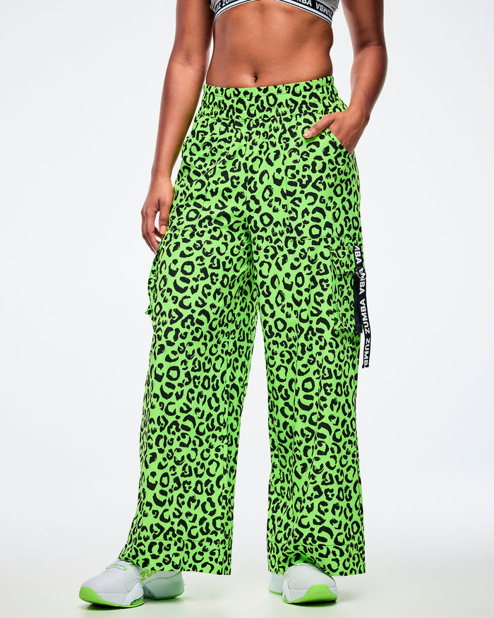 Female model with her left hand in a pocket, of the high-waisted neon green cargo pants with black leopard print, elastic waistband, and side cargo pocket featuring a vertical black tape that reads “Zumba.” Styled with white athletic sneakers accented in matching neon green all on a white background.