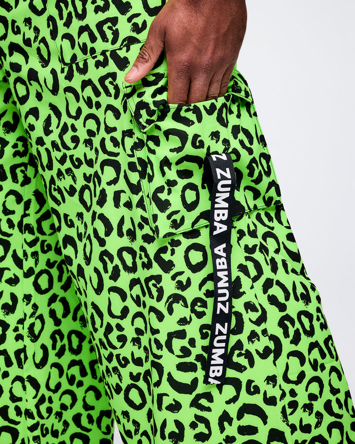 Close-up of neon green leopard-print Zumba cargo pants, hand tucked into the cargo pocket. A vertical black tag along the pocket reads “Zumba" multiple times.
