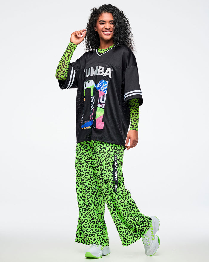 Female model wearing an oversized black Zumba jersey over a neon green leopard-print long-sleeve, paired with matching wide-leg cargo pants featuring a black “Zumba” pocket tape, and white sneakers with neon green accents.