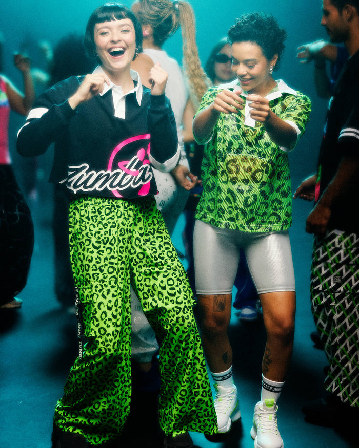 Two female models dancing in a party setting, both wearing matching neon green and black leopard-print outfits. The model on the left wears a black polo with a pink and white Zumba graphic. The model on the right wears a green leopard-print top and silver biker shorts.