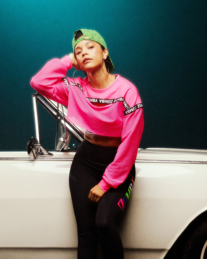 Female model leaning against a white car, wearing a bright pink crop top, black leggings, and a green baseball hat.