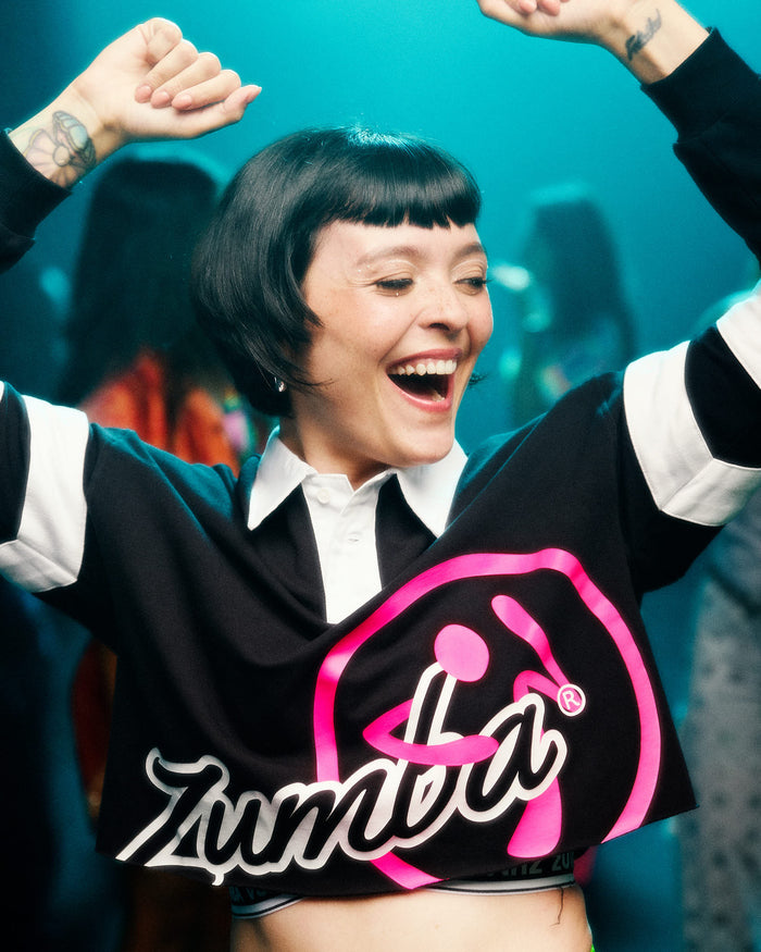 Female model, arms raised dancing in a club, wearing a black cropped polo with white sleeve stripes, a white collar and a large pink and white 'Zumba' logo on the front.
