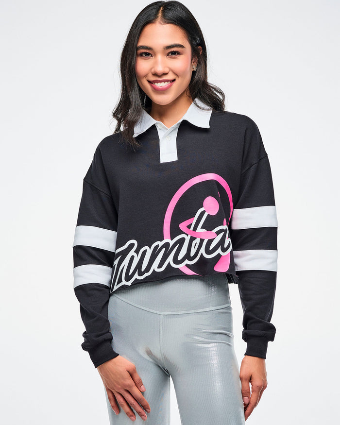 Female model wearing a black cropped polo with white sleeve stripes, a white collar and a large pink and white 'Zumba' logo on the front paired with silver biker shorts.